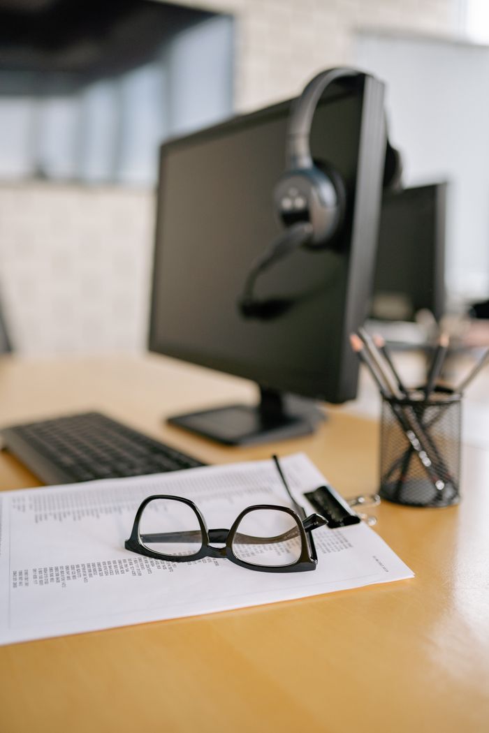 Eyeglasses on a paper next to a computer and headset on a wooden desk.