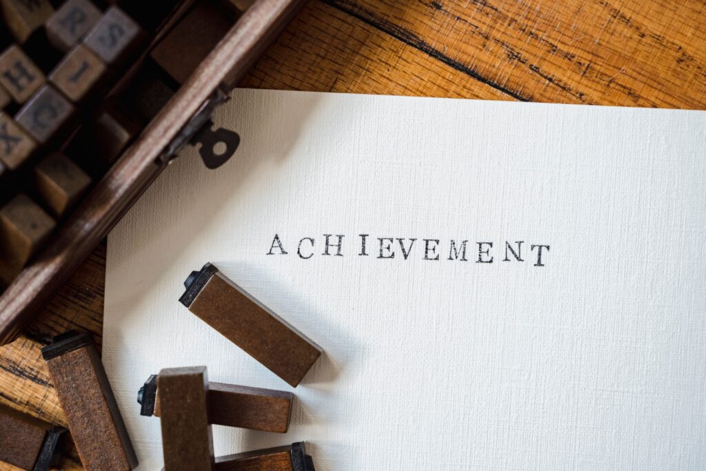 pexels-photo-9017625-9017625 A close-up of wooden stamp typography creating the word 'achievement' on white paper.
